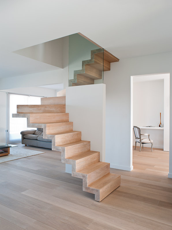 Moderne zwevende trap in licht hout met zigzagprofiel en glazen balustrade, centraal geplaatst in een open woonruimte met houten vloer, witte muren en zicht op een zitkamer en thuiskantoor.