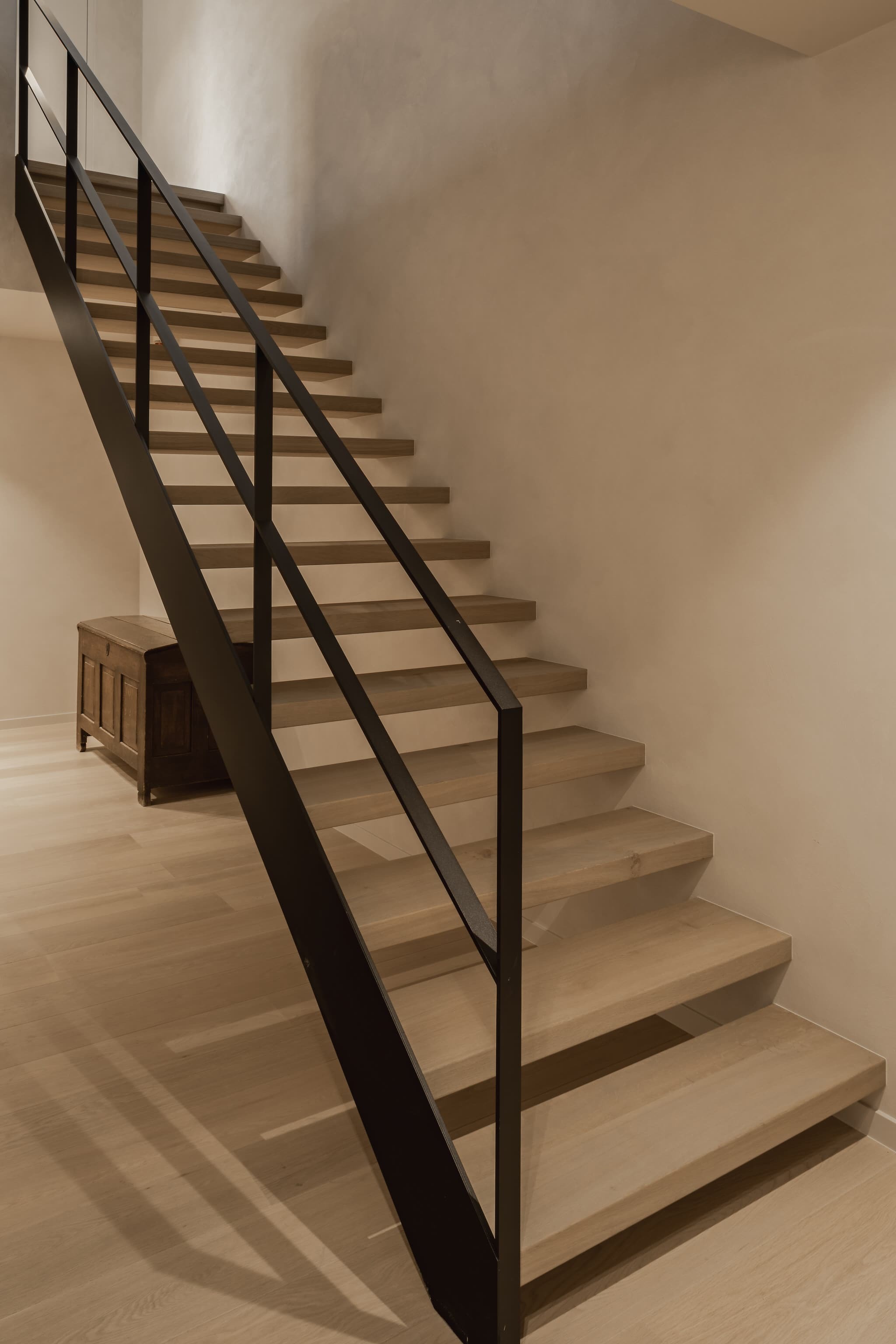 Open houten trap met zwevende treden en een zwarte metalen balustrade, geplaatst in een warm verlichte ruimte met houten vloer en een houten kast tegen de muur.