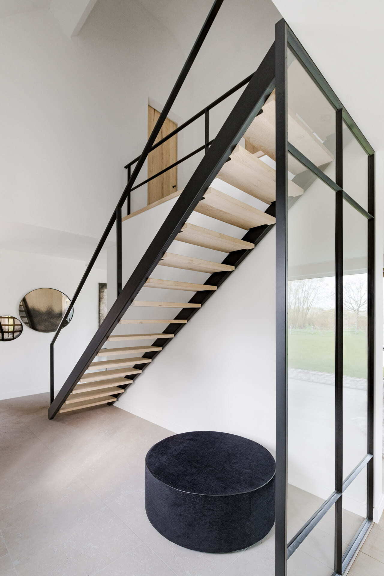Moderne zwevende trap met zwarte leuning en houten treden in een licht interieur, met een zwarte poef op de voorgrond en een glazen wand rechts.